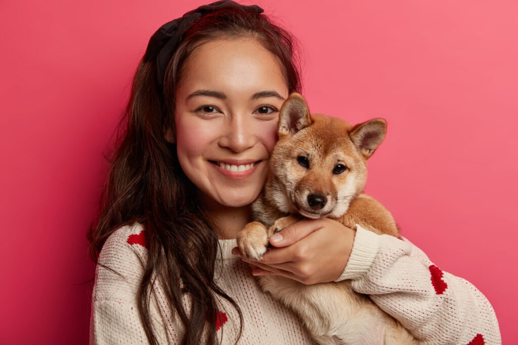 Headshot of cheerful brunette young woman adopts small puppy, embraces pedigree dog, smiles pleasantly, cares about pet, wears warm jumper, isolated on pink background, have walk together at weekend.